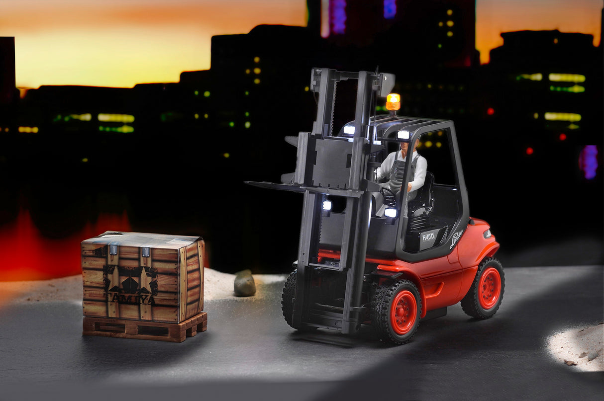 The Carson 1/14 Linde RC Forklift navigates a city landscape at sunset, lifting a crate of cargo against a backdrop of skyscrapers.