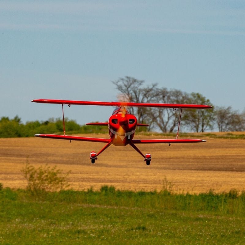 Red and black remote-controlled biplane model flying over a grassy field with trees in the background.