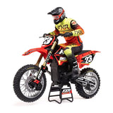 Detailed RC motorcycle racer in FXR outfit, riding Losi Promoto-MX 1/4 scale dirt bike on track.