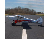 Sleek gray and red Hangar 9 Cub Crafters XCub 60cc ARF RC Plane on asphalt runway with natural surroundings.