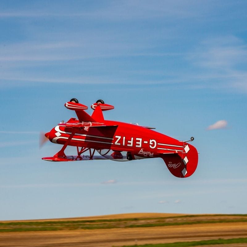 Bright red Hangar 9 Pitts S-2B 60cc ARF model aircraft soaring against a clear blue sky over an open field.