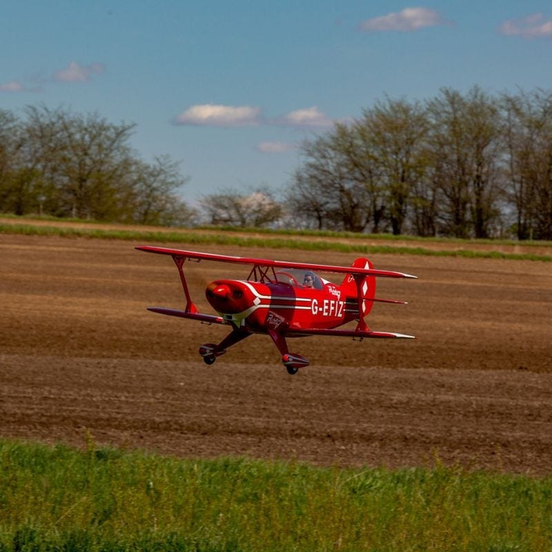 Vibrant red Hangar 9 Pitts S-2B 60cc ARF RC plane soaring across grassy field against blue sky with clouds