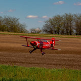 Vibrant red Hangar 9 Pitts S-2B 60cc ARF RC plane soaring across grassy field against blue sky with clouds